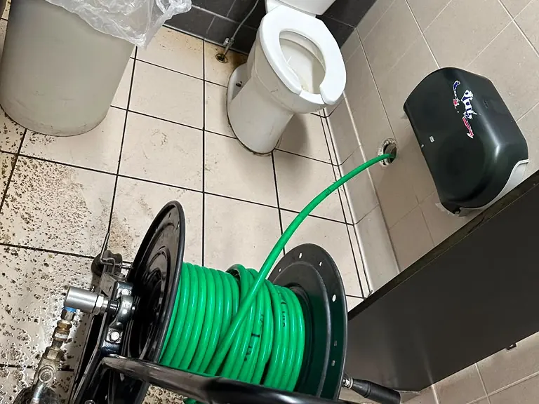 Hydro jetter hose feeding into a commercial restroom floor cleanout beside the toilet — hydro jetting in Bergenfield, NJ
